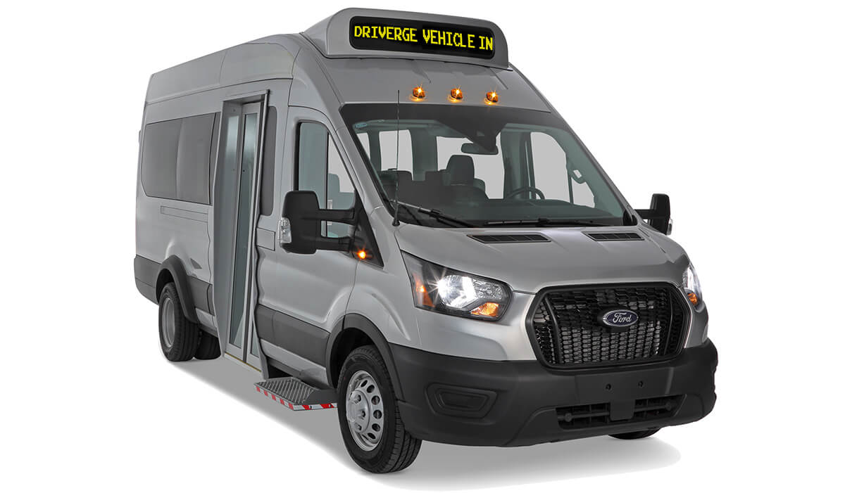 Un minibus Ford argenté servant de navette pour passagers, avec une porte d'entrée avant et un panneau numérique suspendu indiquant « CONDUCTEUR VÉHICULE ENTRÉE ».
